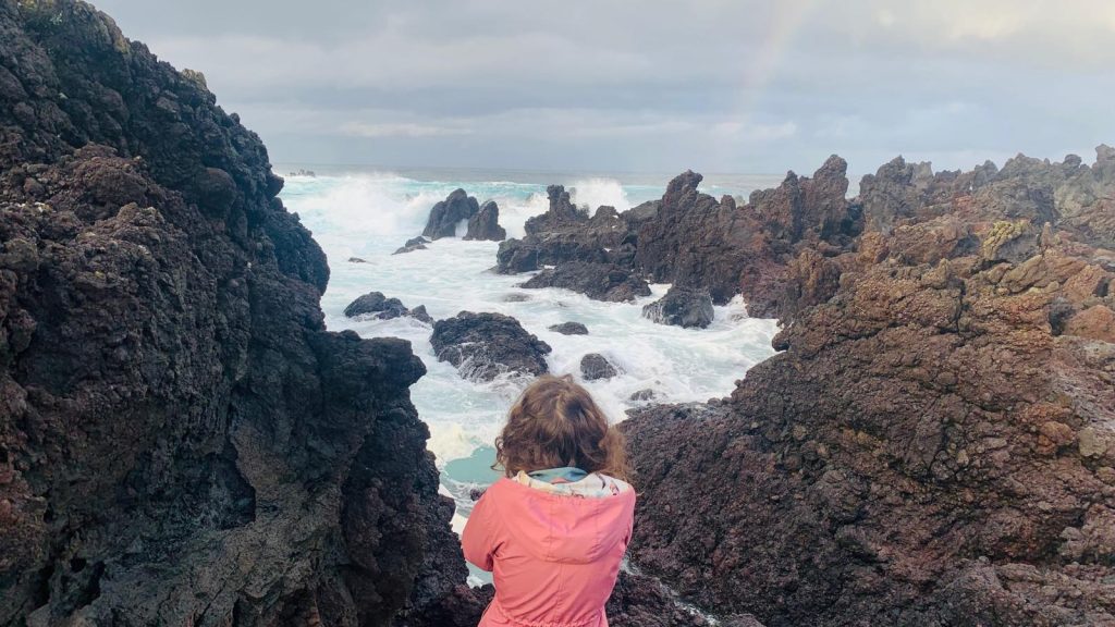 Bild bei dem eine Frau im rosa Mantel an einer Felsenküste sitzt und den Wellen zuschaut.